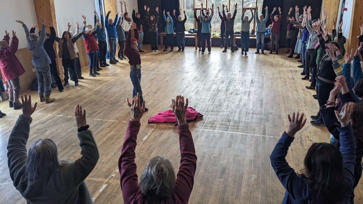 A group of people standing in a large circle in a bright hall, doing a group warm up before singing. All the people have their arms in the air